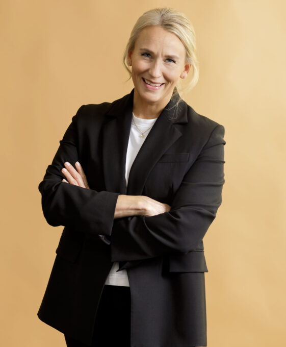 A person with blond hair in a black suit and white shirt stands smiling with arms crossed against a light tan background.