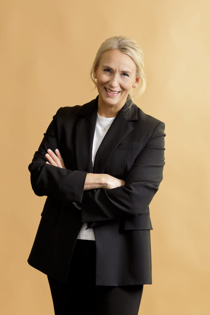 A person with blond hair in a black suit and white shirt stands smiling with arms crossed against a light tan background.