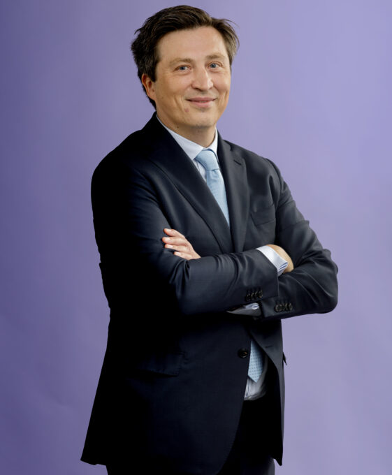 A man in a dark suit and light blue tie stands with arms crossed, smiling, in front of a plain purple background.