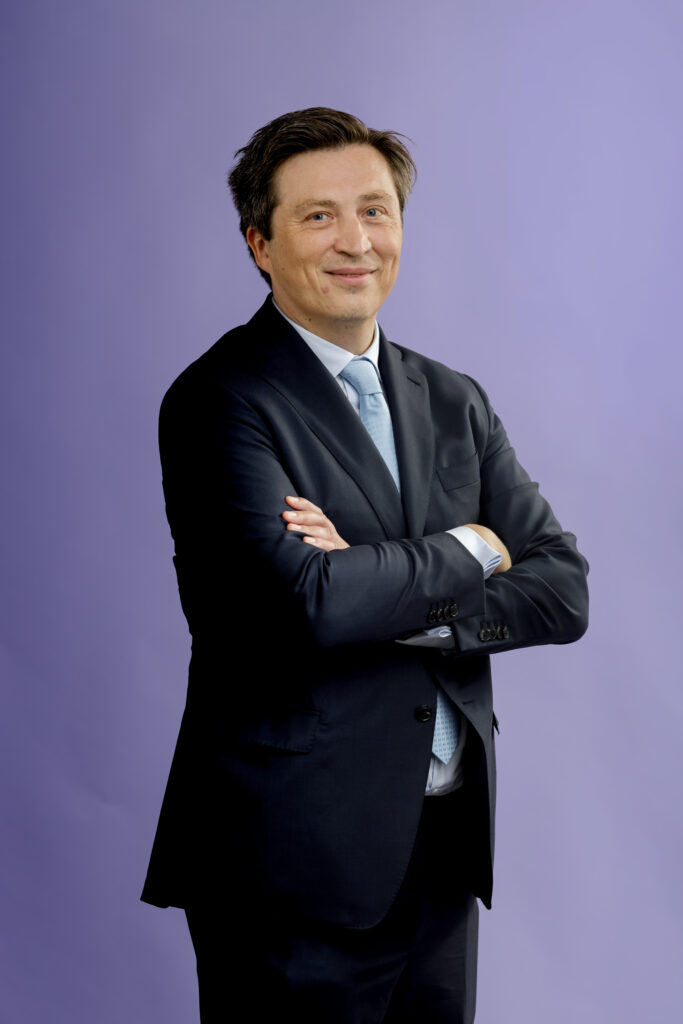 A man in a dark suit and light blue tie stands with arms crossed, smiling, in front of a plain purple background.