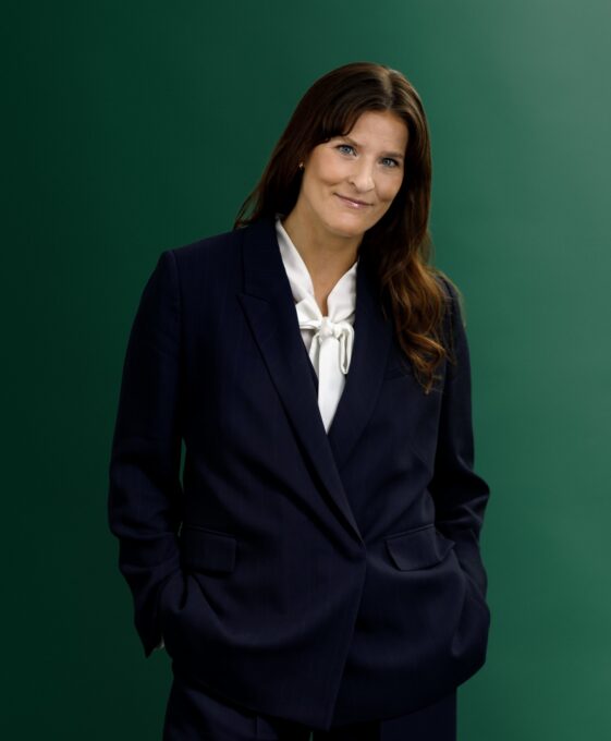 A woman wearing a navy blue suit and white blouse stands against a green background, with her hands in her pockets and a slight smile on her face.