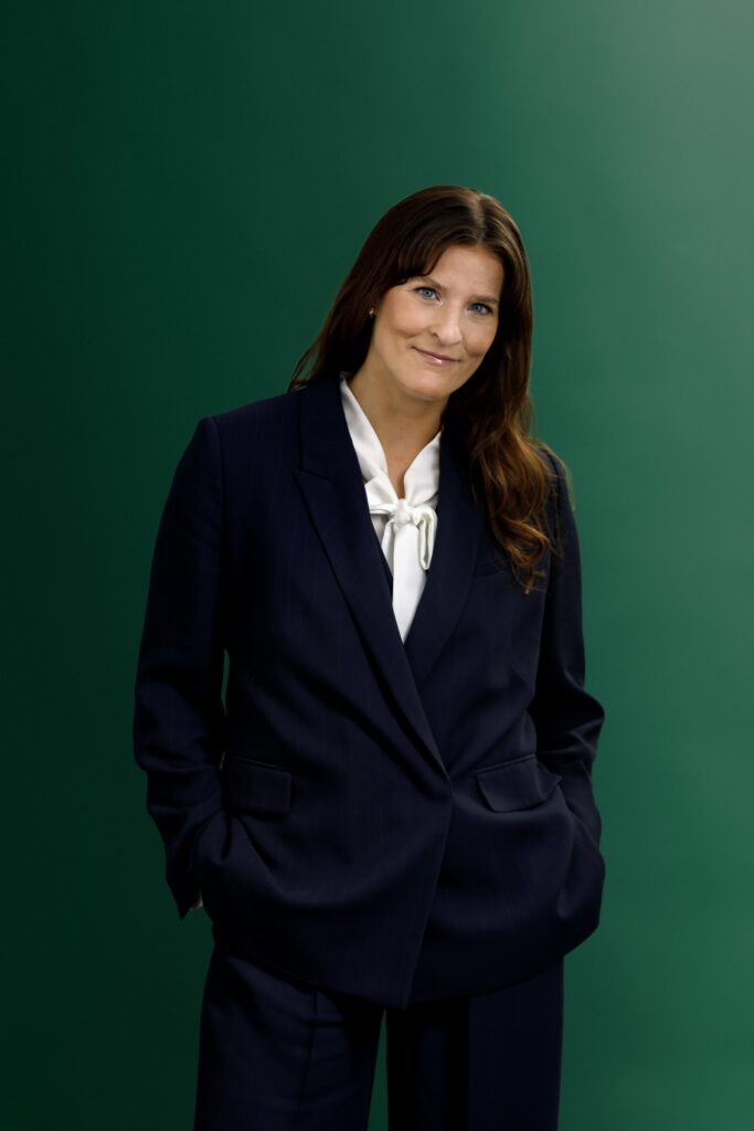 A woman wearing a navy blue suit and white blouse stands against a green background, with her hands in her pockets and a slight smile on her face.