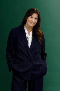 A woman wearing a navy blue suit and white blouse stands against a green background, with her hands in her pockets and a slight smile on her face.