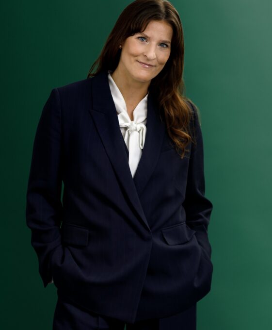 A woman wearing a navy blue suit and white blouse stands against a green background, with her hands in her pockets and a slight smile on her face.