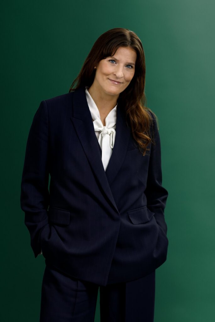 A woman wearing a navy blue suit and white blouse stands against a green background, with her hands in her pockets and a slight smile on her face.
