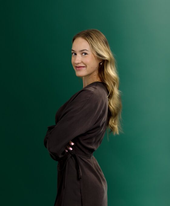 A woman with long, wavy blonde hair in a brown dress stands sideways with arms crossed, smiling against a plain green background.