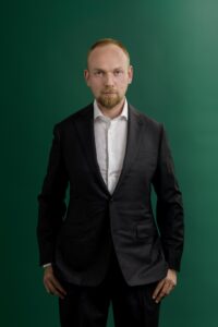A man in a dark suit and white shirt stands against a green background, looking directly at the camera with a neutral expression and his hands by his sides.