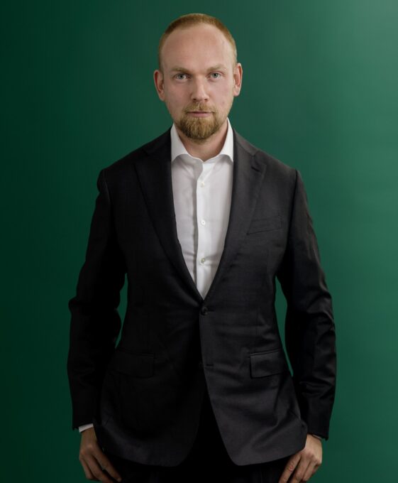 A man in a dark suit and white shirt stands against a green background, looking directly at the camera with a neutral expression and his hands by his sides.