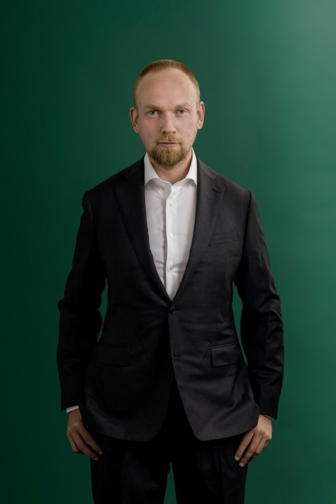 A man in a dark suit and white shirt stands against a green background, looking directly at the camera with a neutral expression and his hands by his sides.