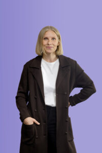 A woman wearing a dark coat over a white t-shirt stands against a purple background.