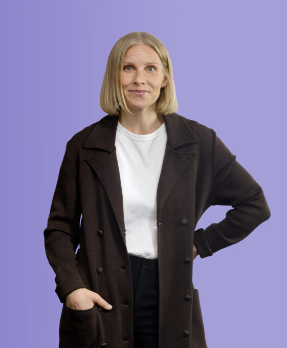 A woman wearing a dark coat over a white t-shirt stands against a purple background.