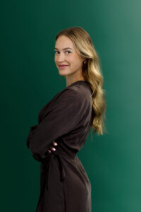 A woman with long, wavy blonde hair stands in profile against a green background, wearing a dark wrap dress and smiling slightly at the camera.