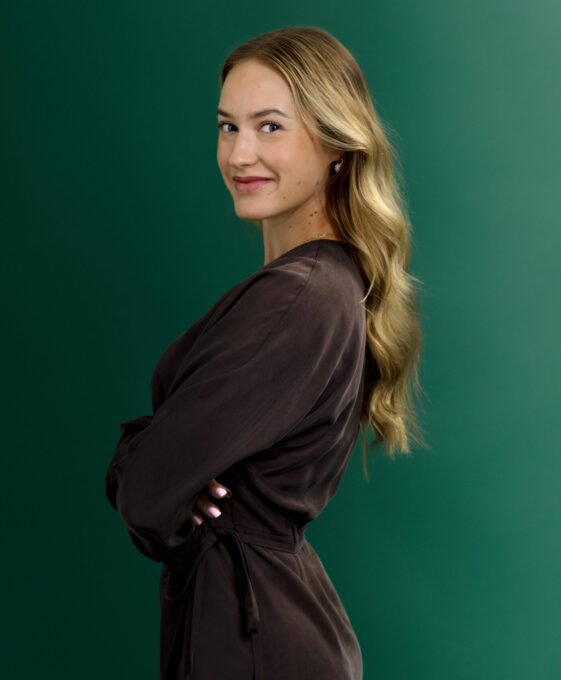 A woman with long, wavy blonde hair stands in profile against a green background, wearing a dark wrap dress and smiling slightly at the camera.