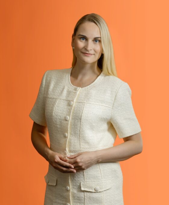 A woman with blonde hair wearing a cream-colored, short-sleeved button-up jacket stands against an orange background.