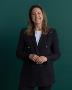A woman with light brown hair wearing a dark blazer and white shirt stands smiling against a solid green background. She has her hands gently clasped in front of her.