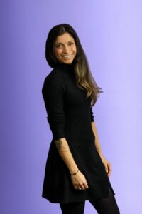 A woman with long dark hair wearing a black long-sleeve top and skirt stands smiling against a light purple background. She has a geometric tattoo on her forearm.
