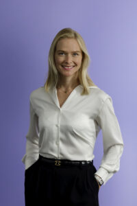 A woman with blonde hair wearing a white blouse and black pants stands against a light purple background, smiling with her hands in her pockets.