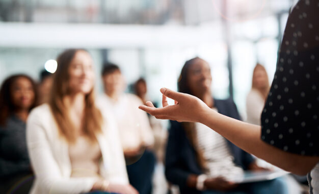 A person stands in the foreground, gesturing with their hand while speaking to a blurred audience seated indoors, suggesting a presentation or seminar setting.