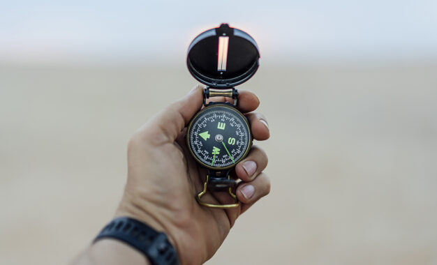 A person’s hand holding an open compass, pointing north, against a blurred outdoor background. The person is wearing a dark wristwatch.