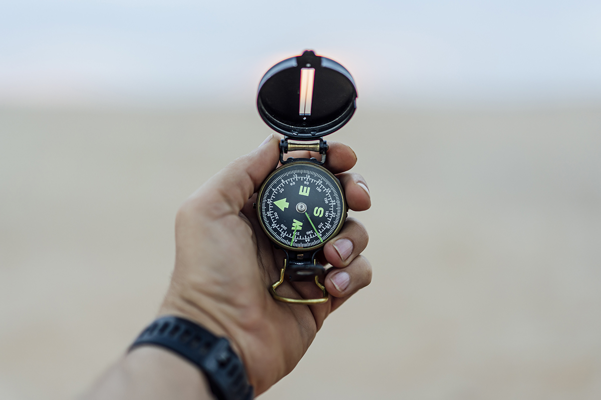 A person’s hand holding an open compass, pointing north, against a blurred outdoor background. The person is wearing a dark wristwatch.