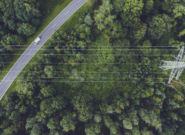 Aerial view of a car driving on a road through a forest, with power lines and a transmission tower crossing above the green trees.