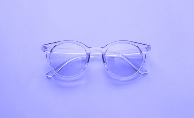 Clear round eyeglasses with transparent frames are placed on a smooth, light purple surface, photographed from above.