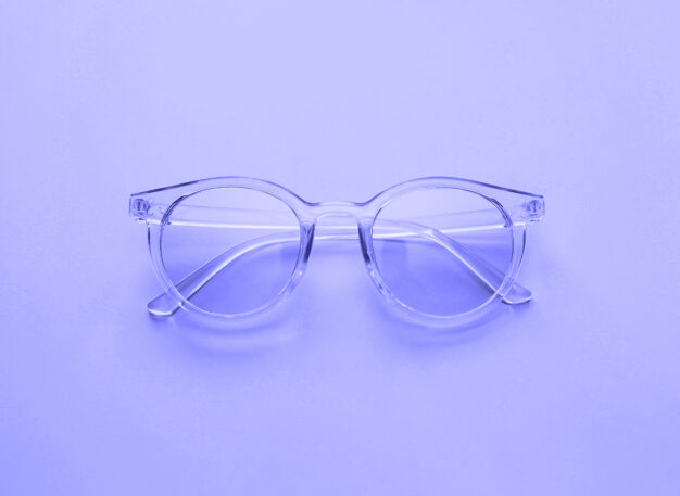 Clear round eyeglasses with transparent frames are placed on a smooth, light purple surface, photographed from above.