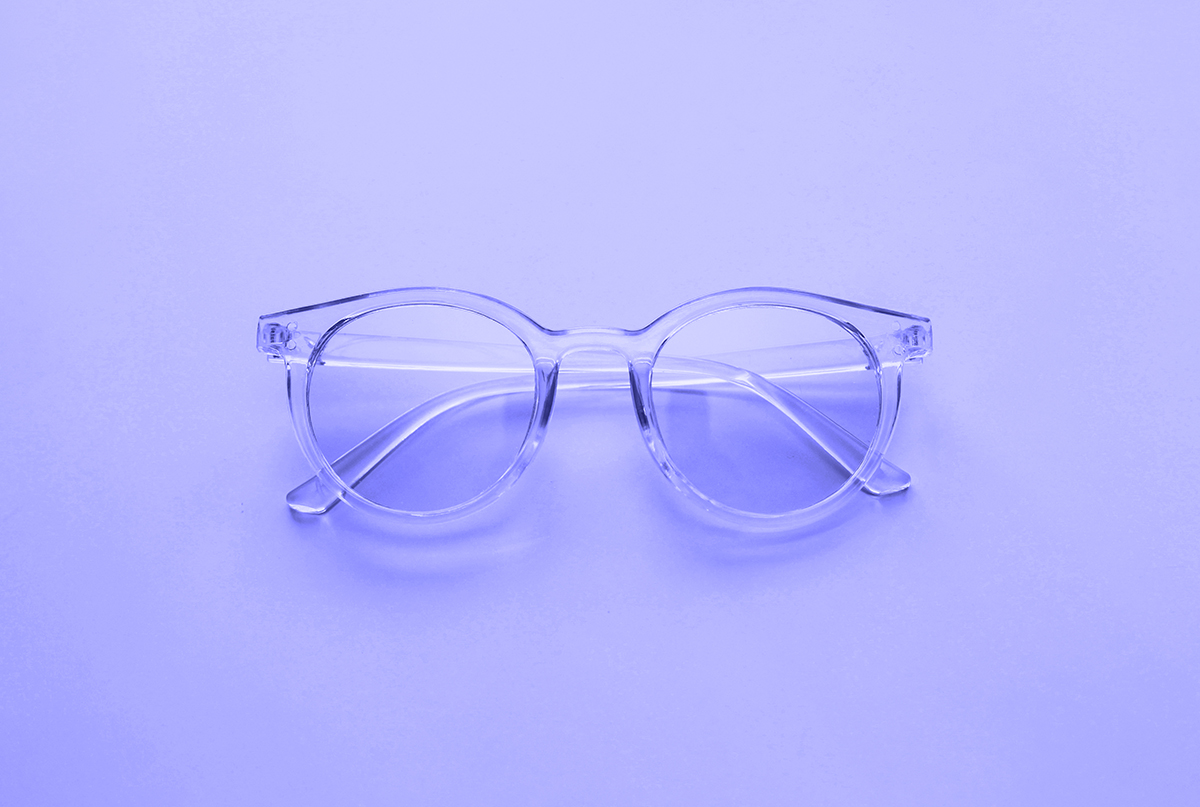 Clear round eyeglasses with transparent frames are placed on a smooth, light purple surface, photographed from above.