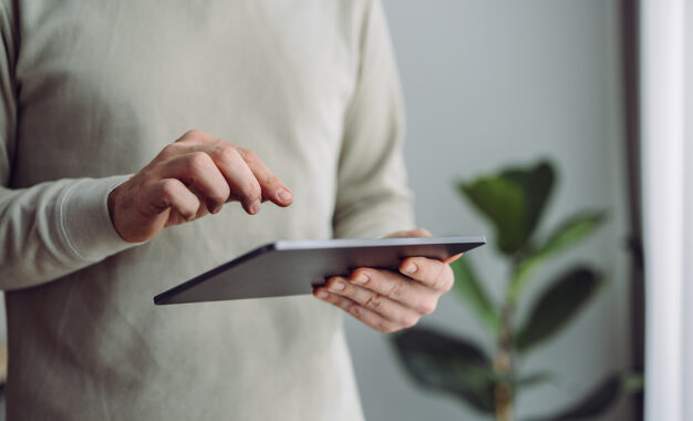 A person wearing a light-colored long-sleeve shirt uses both hands to operate a tablet. A leafy green plant and a softly lit window are visible in the blurred background.