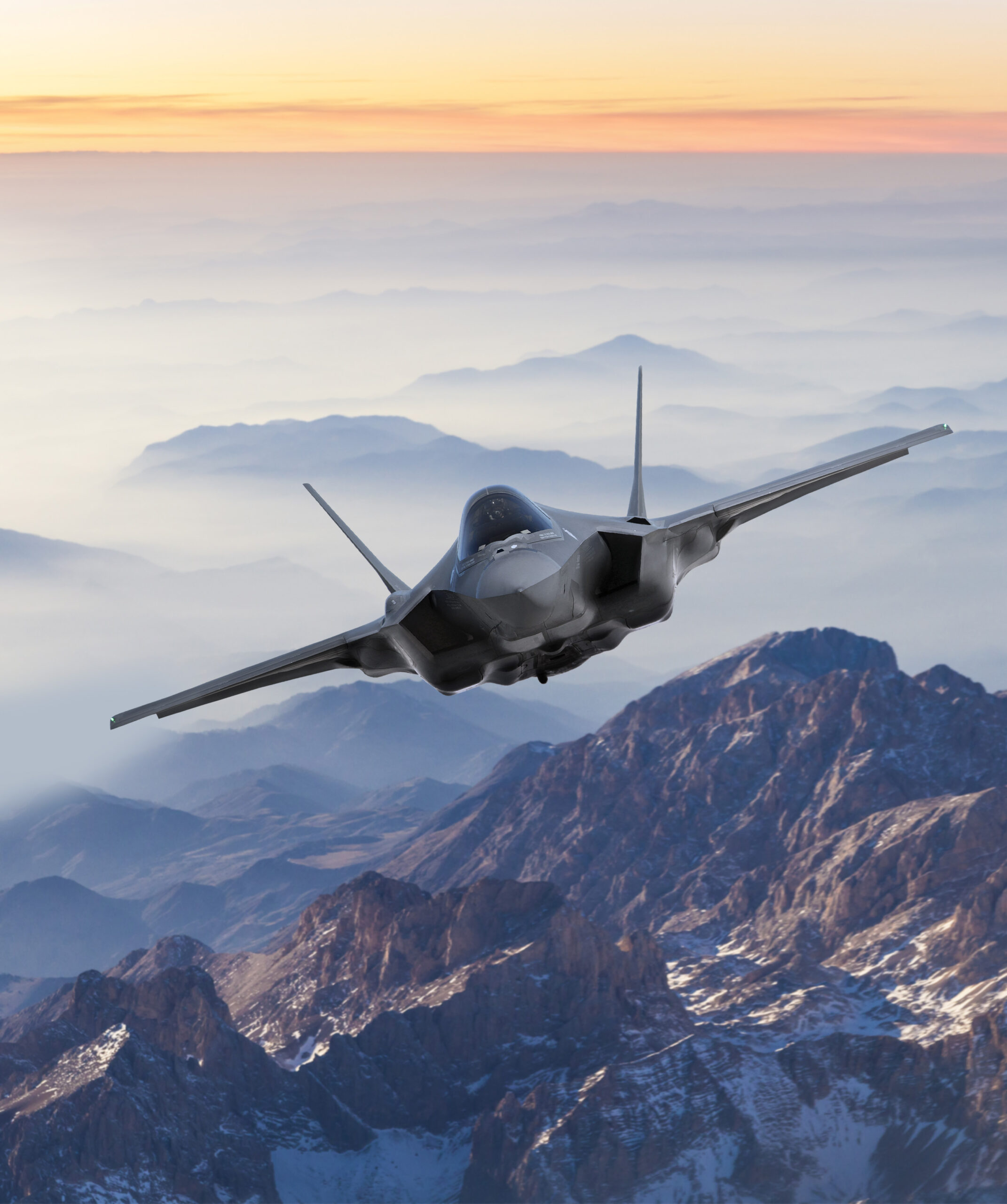 A stealth fighter jet flies above rugged, snow-dusted mountains at sunrise, with layers of mist and a colorful sky in the background.