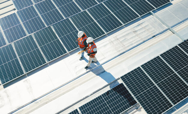 Two workers in safety gear and orange vests walk between rows of solar panels on a rooftop, inspecting the installation under bright sunlight.