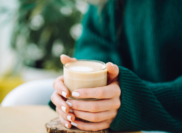 A person in a dark green sweater holds a glass mug of cappuccino or latte with both hands, sitting at a light-colored table. The background is blurred with some greenery visible.