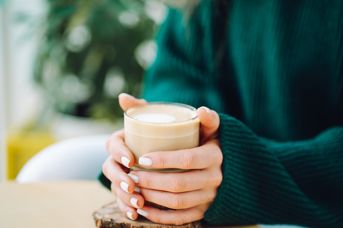 A person in a dark green sweater holds a glass mug of cappuccino or latte with both hands, sitting at a light-colored table. The background is blurred with some greenery visible.