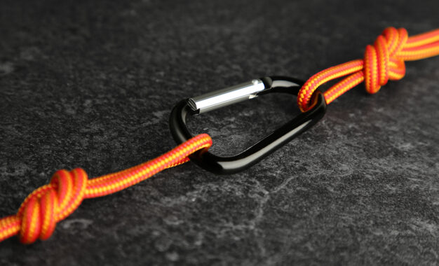 A black carabiner connects two orange ropes with knots on a dark textured surface.
