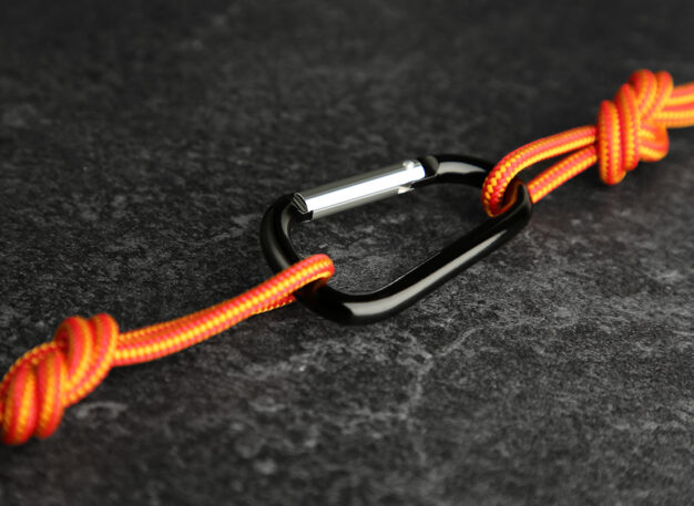 A black carabiner connects two orange ropes with knots on a dark textured surface.