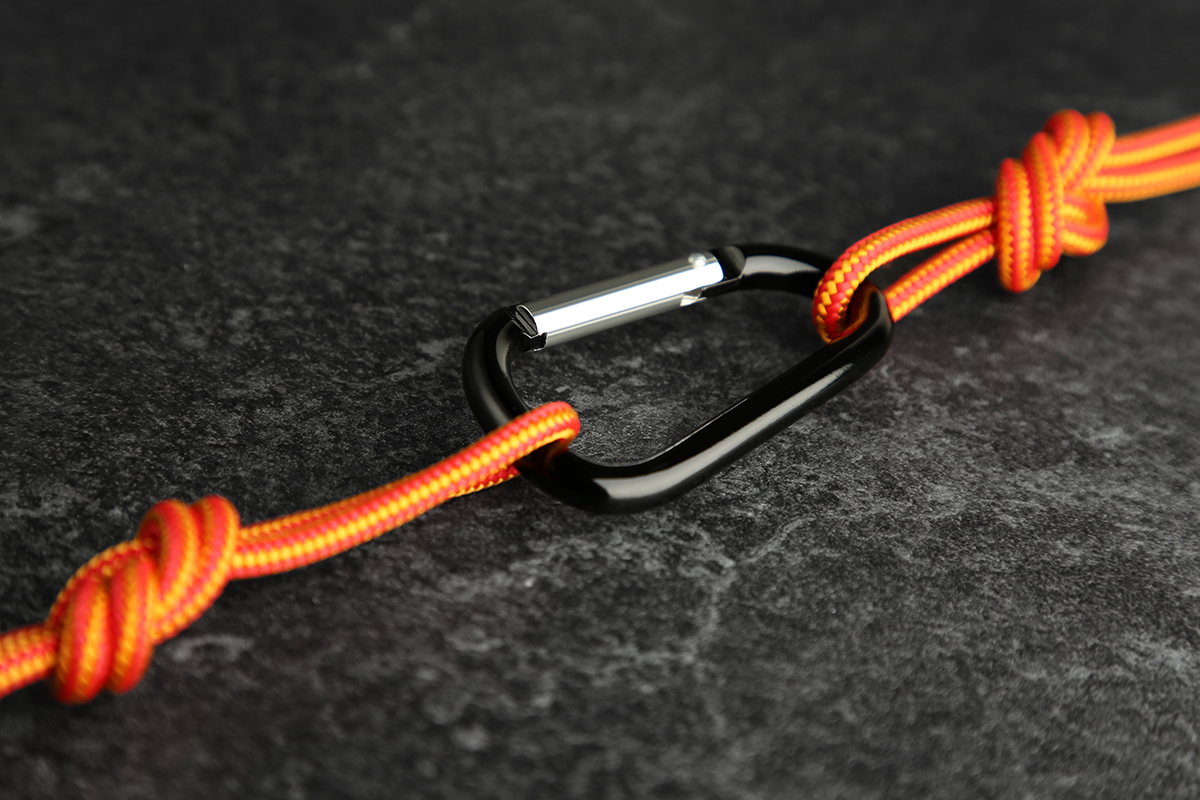 A black carabiner connects two orange ropes with knots on a dark textured surface.