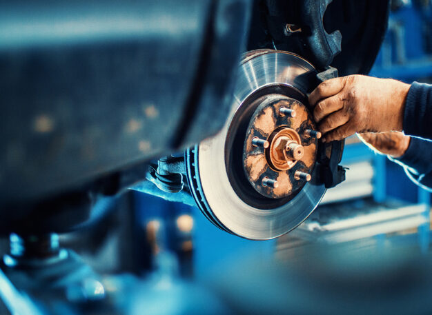 A mechanic’s hands work on a car’s exposed disc brake system in a garage, adjusting parts with tools. The background is blurred, highlighting the focus on the vehicle repair.