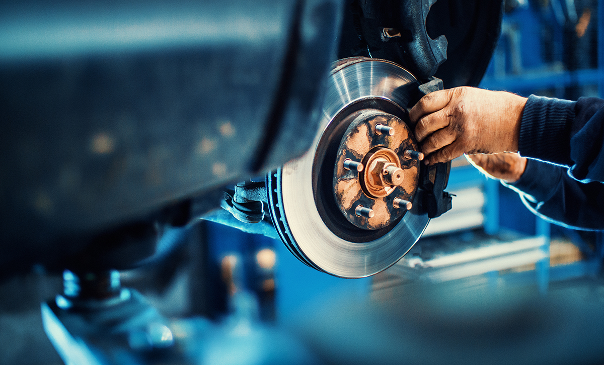 A mechanic’s hands work on a car’s exposed disc brake system in a garage, adjusting parts with tools. The background is blurred, highlighting the focus on the vehicle repair.
