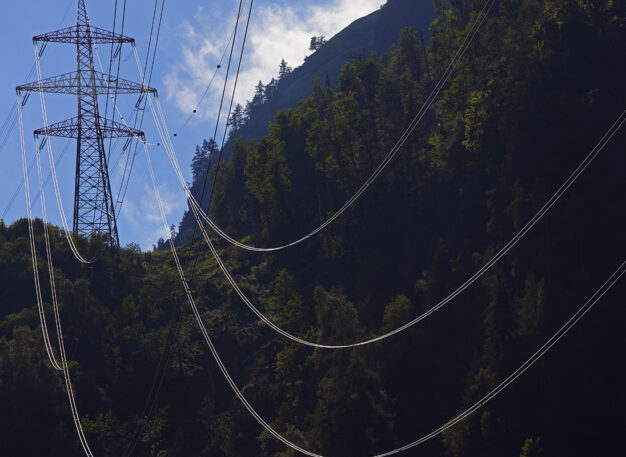 Tall electricity transmission towers and power lines stretch across a forested hillside under a partly cloudy sky. Sunlight highlights the wires and some tree tops, while much of the forest remains in shadow.