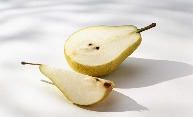 A ripe yellow pear sliced in half, with one half lying flat and the other piece, a quarter segment, resting in front on a white surface with soft shadows.