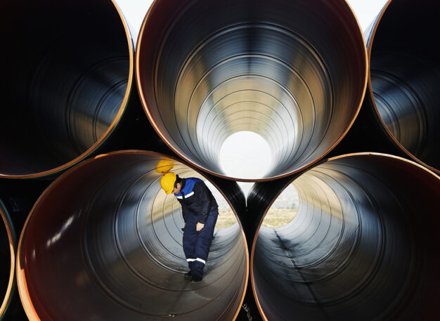 A worker wearing a yellow hard hat and blue uniform inspects the inside of a large metal pipe, surrounded by stacked, hollow steel pipes in an industrial setting.