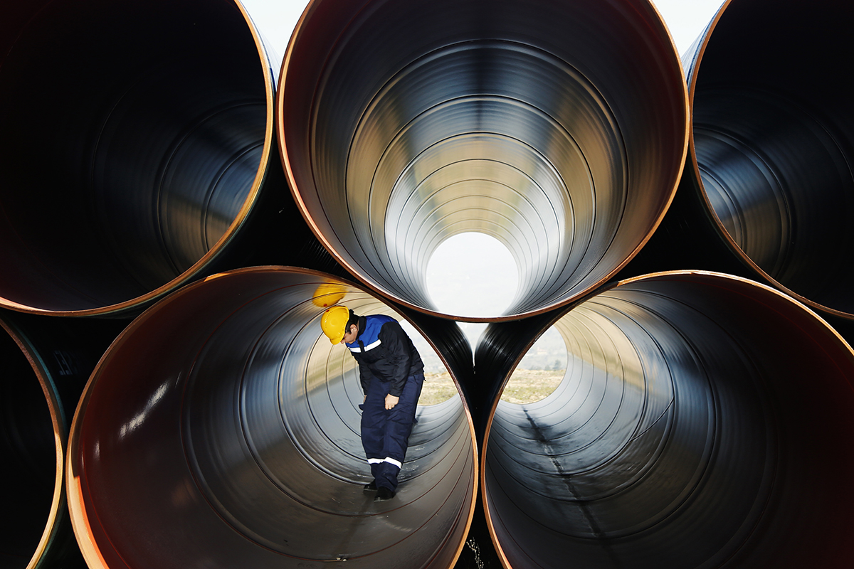 A worker wearing a yellow hard hat and blue uniform inspects the inside of a large metal pipe, surrounded by stacked, hollow steel pipes in an industrial setting.