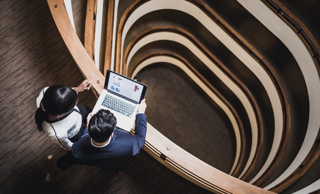 Two business professionals view a laptop displaying charts while standing near the railing of a modern, multi-story building with a spiraling interior design seen from above.