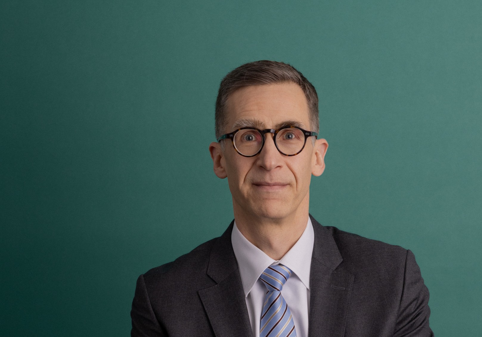 A man wearing glasses and a dark suit with a light blue striped tie sits in front of a plain green background, facing the camera with a neutral expression.