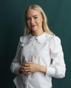 A woman with long blonde hair stands in front of a dark green background, wearing a white blouse with a ruffled collar and smiling gently with her hands clasped together.