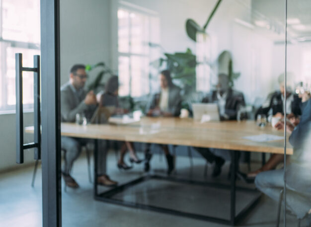 A group of people in business attire sit around a conference table in a modern office, seen through a glass door, engaged in a meeting with laptops and papers on the table.