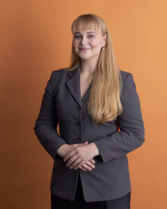 A woman with long blonde hair wearing a gray blazer stands smiling with hands folded in front of an orange background.