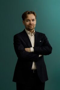 A man with light brown hair and a beard stands against a teal background, wearing a dark suit and a light-colored dress shirt, with his arms crossed and a slight smile on his face.