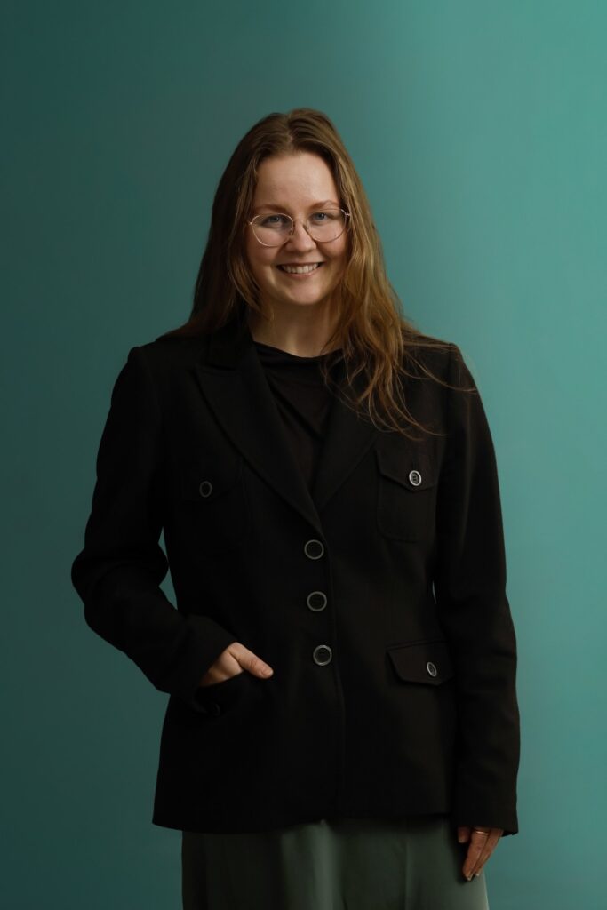 A woman with long brown hair and glasses smiles at the camera. She is wearing a black blazer over a dark top, with one hand in her pocket, standing in front of a teal background.