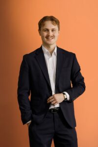 A young man with light brown hair, wearing a dark suit and white dress shirt, stands smiling against an orange background. One hand is in his pocket while the other rests near his jacket button.